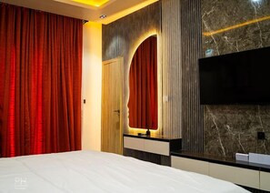 A modern bedroom with a large bed, a TV mounted on the wall, and a vanity with a mirror. - Palatial Apartments  (Lekki)