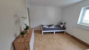 Una habitación pequeña y luminosa con piso de madera, una cama blanca con colcha floral, una mesita con una planta en maceta y una ventana que proporciona luz natural.
