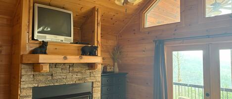 A cozy cabin interior with a stone fireplace, wooden shelves, a television, and a ceiling fan.