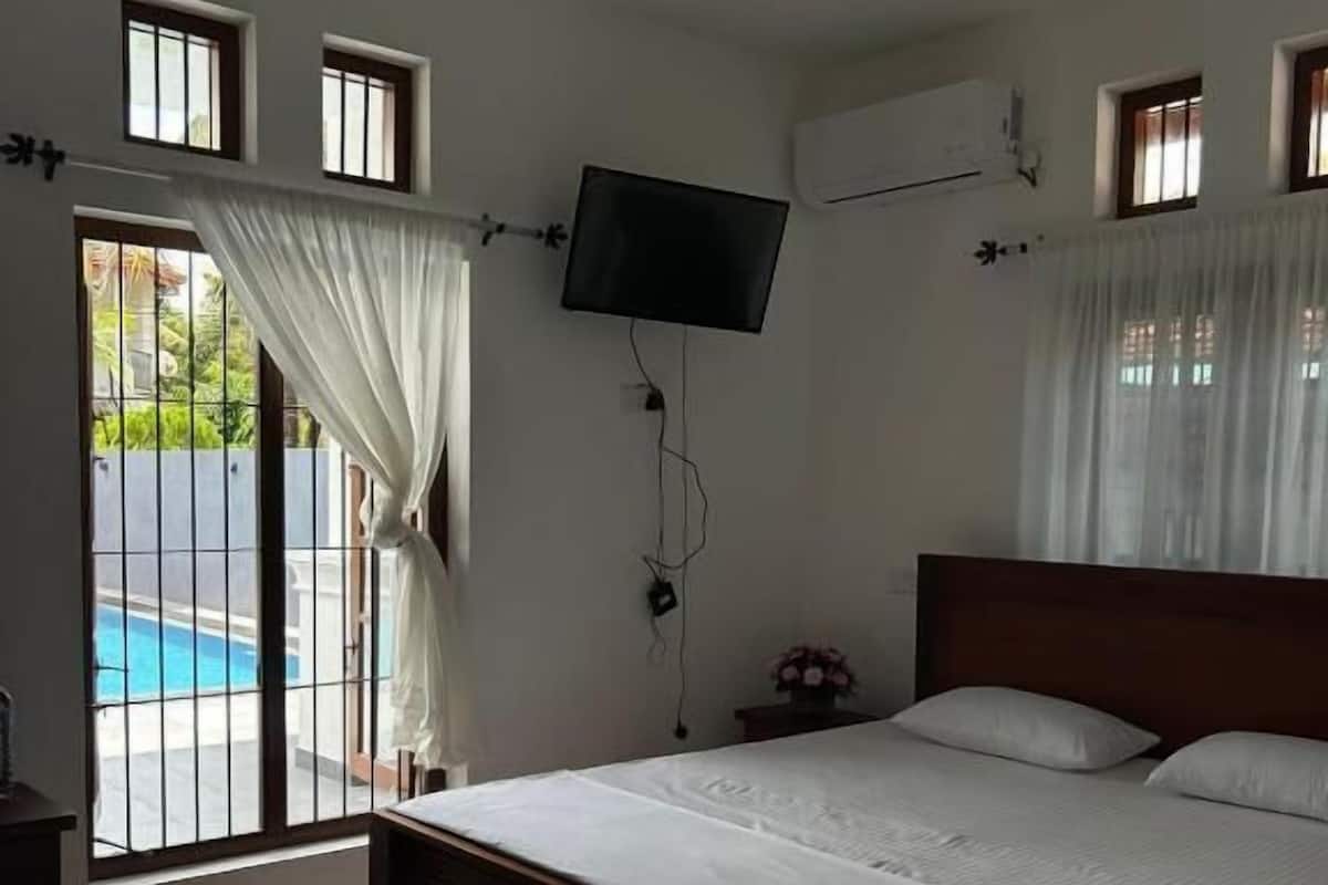 Quarto com cama, ventilador de teto, televisão na parede e vista para uma piscina pela janela.