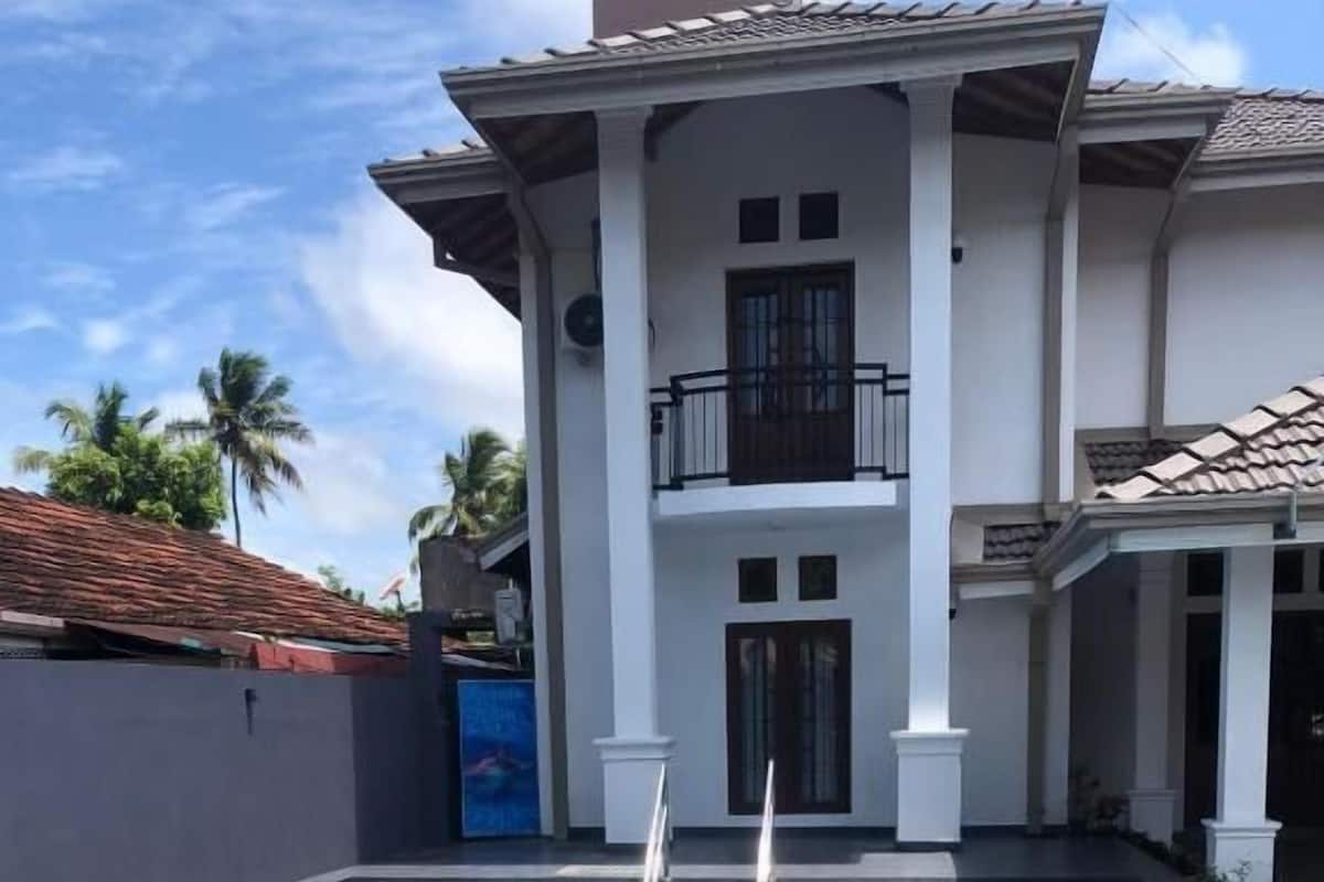 Uma piscina com deck ladrilhado, um edifício de dois andares com varanda e telhado de telhas vermelhas.