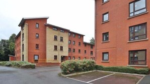 Un immeuble résidentiel à plusieurs étages, doté d’une façade en briques, de nombreuses fenêtres et d’un espace de stationnement agrémenté d’un petit jardin.