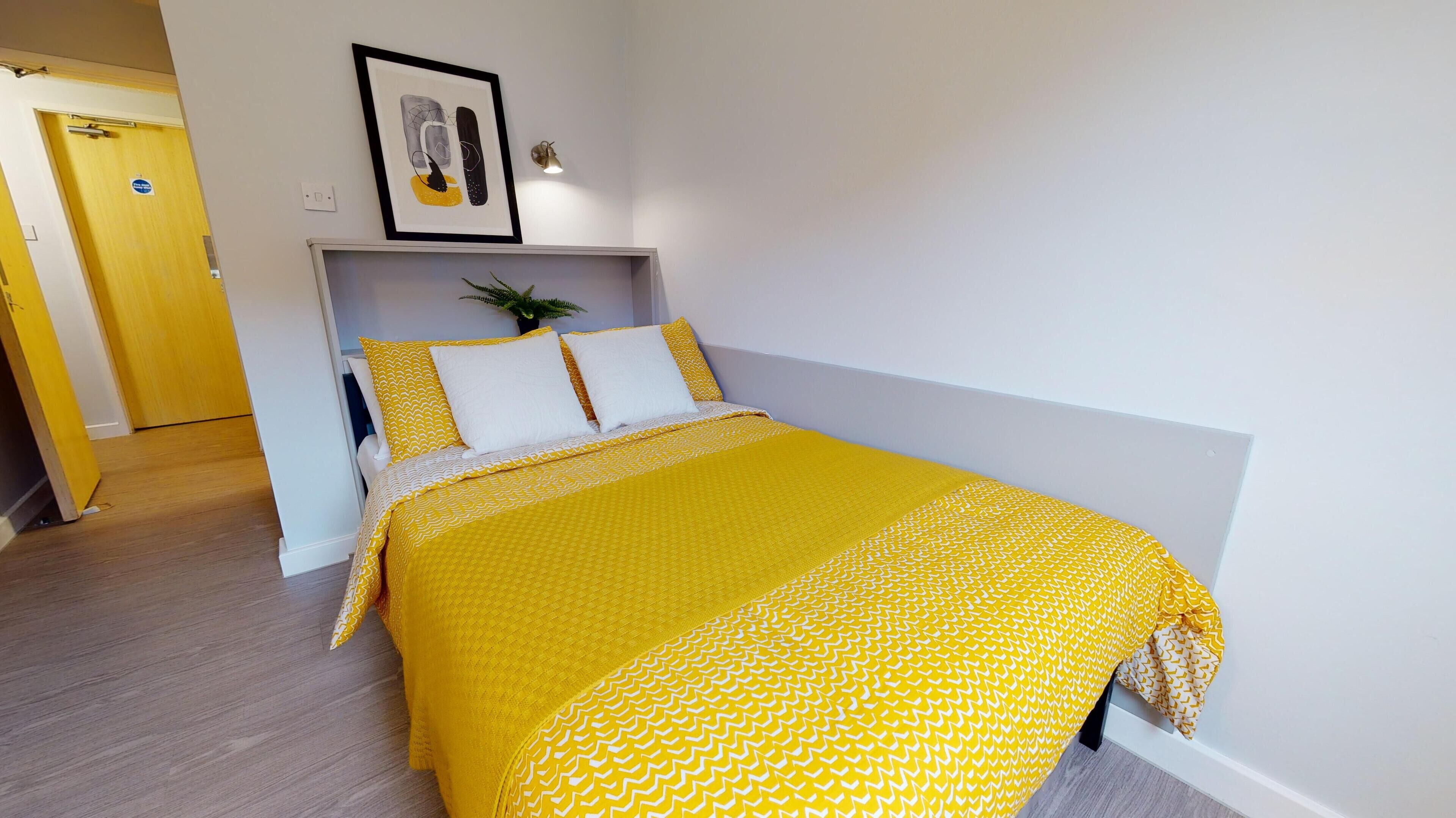 A neatly made bed with a yellow patterned bedspread and pillows, a framed picture above the bed, and a small potted plant on the nightstand.