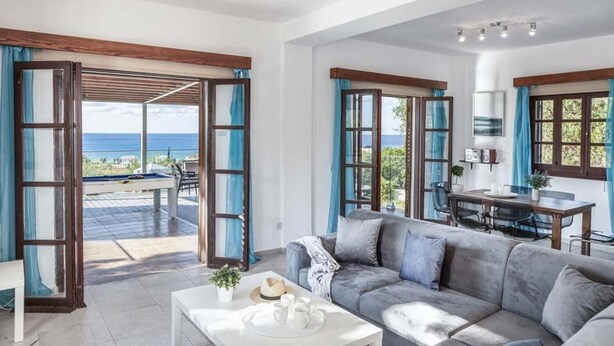 A spacious living area with a grey sofa, a dining table, and a view of the sea through large windows. - Villa Argaka Vista - Three Bedroom Villa, Sleeps 6 (Argaka)