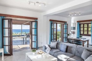 A spacious living area with a grey sofa, a dining table, and a view of the sea through large windows. - Villa Argaka Vista - Three Bedroom Villa, Sleeps 6 (Argaka)