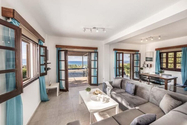 A spacious living room with a large gray sofa, a white coffee table, and a dining area with a table and chairs. There are large windows with blue curtains, offering a view of the outdoors. - Villa Argaka Vista - Three Bedroom Villa, Sleeps 6 (Argaka)