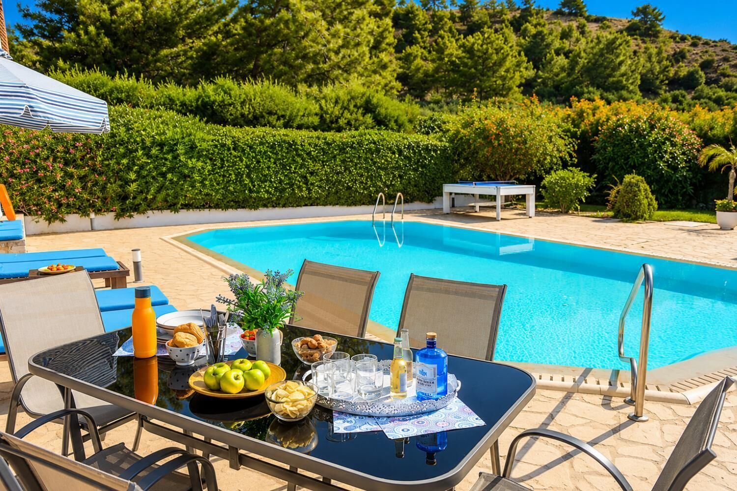 Une table au bord de la piscine, avec de la nourriture et des boissons, entourée de chaises longues et donnant sur la piscine.