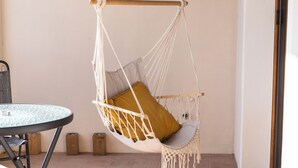 Um quarto com uma cadeira de macramé branca, um assento de almofada amarelo mostarda, uma mesa de vidro redonda e um banquinho de madeira.