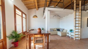 Um quarto amplo com uma mesa de jantar de madeira, cadeiras e um vaso com planta. Há um sofá e uma escada ao fundo.
