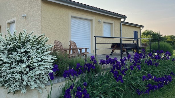 Un bâtiment à l’extérieur de couleur beige, deux chaises en bois, une table en métal et, au premier plan, des fleurs violettes.