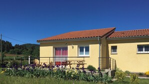 Un bâtiment jaune avec un toit recouvert de tuiles rouges, un banc en bois et, au premier plan, des fleurs violettes et jaunes.