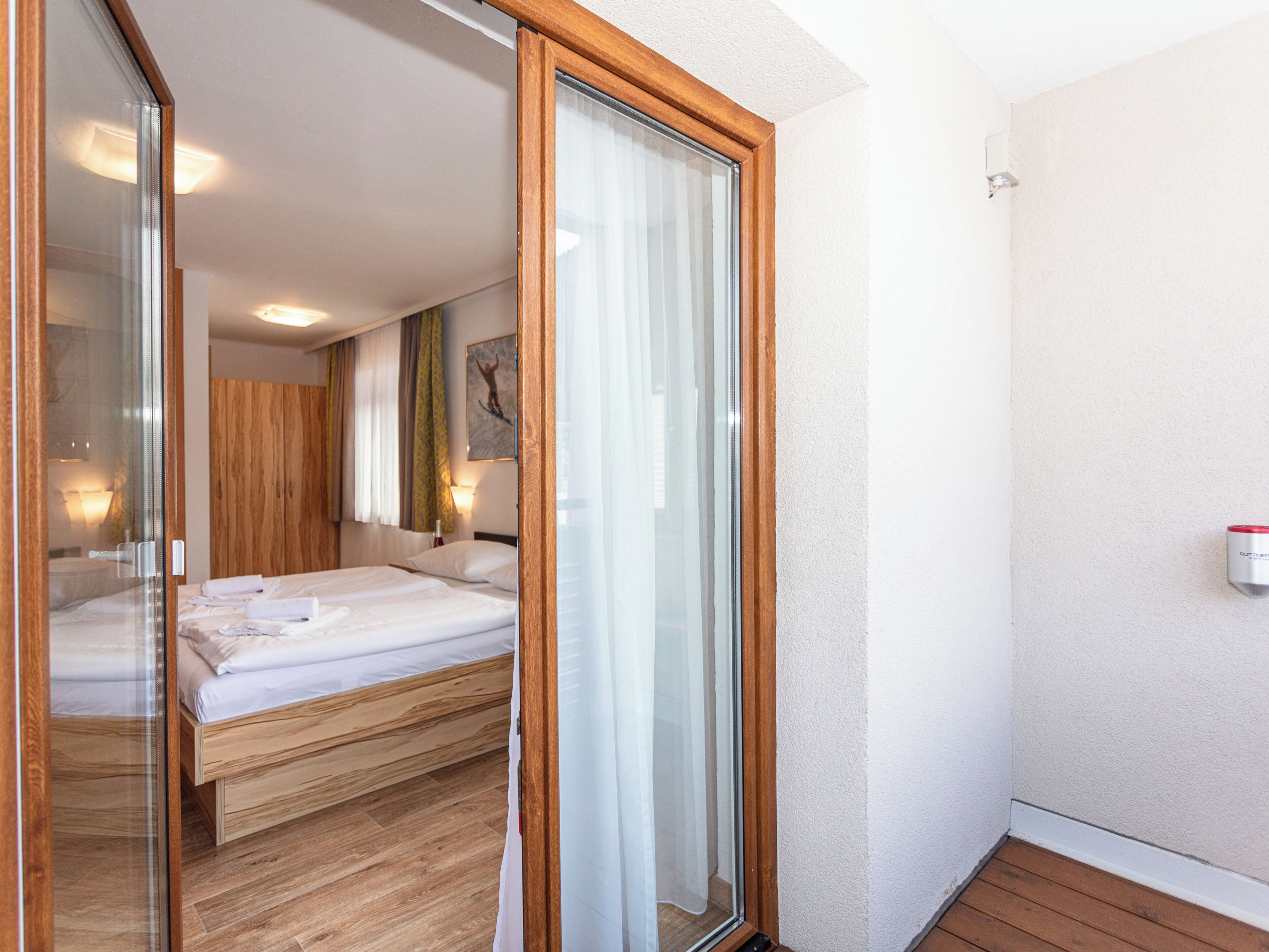 A hotel room with a bed, a sliding glass door, a wooden floor, and a wall-mounted light fixture.