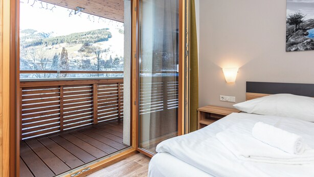 Una camera da letto con pavimento in legno, un letto con lenzuola bianche, un comodino e vista su un paesaggio innevato attraverso una porta in vetro.