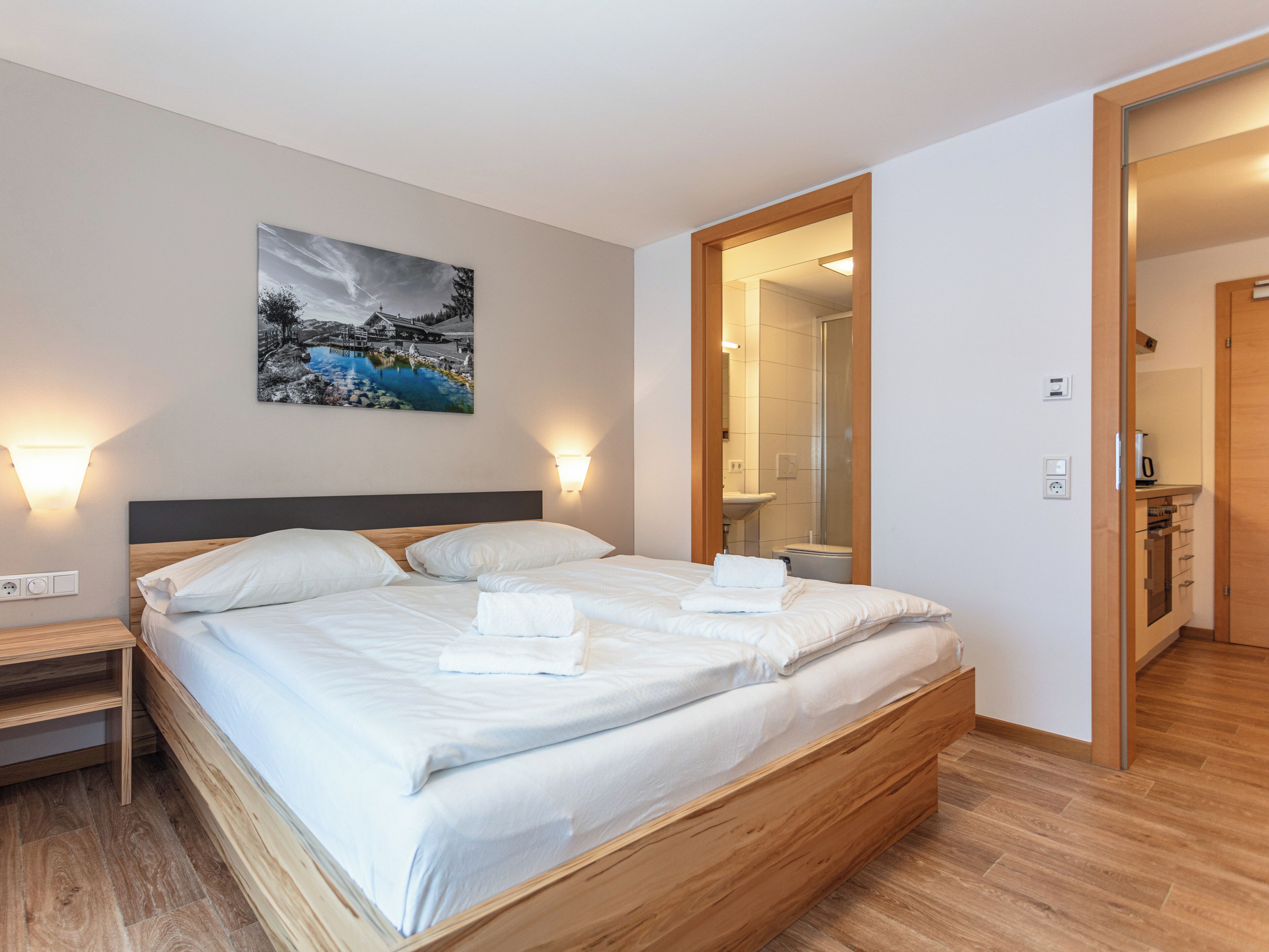 Moderni hotellihuone, jossa on suuri sänky, puinen pääty, maisemamaalaus ja kylpyhuone, joka näkyy avoimesta ovesta.