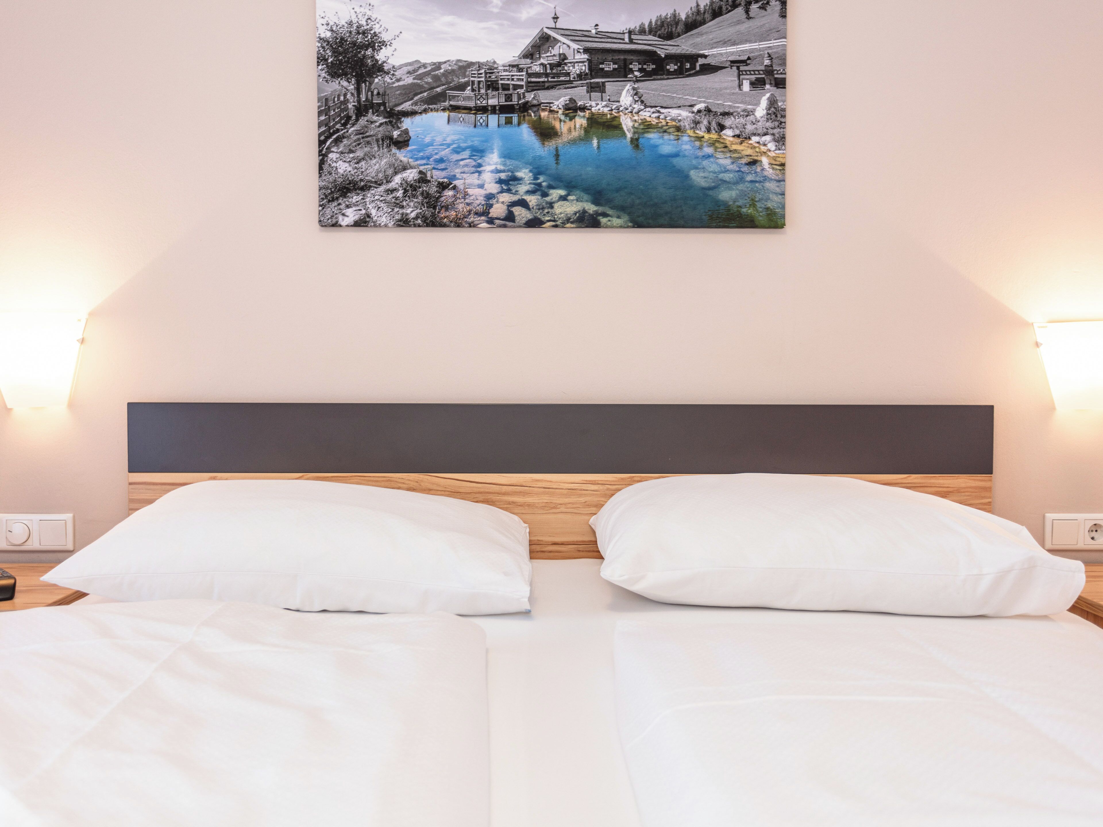 A hotel room with a bed, two white pillows, a wooden headboard, and a wall-mounted picture of a mountain landscape with a lake.