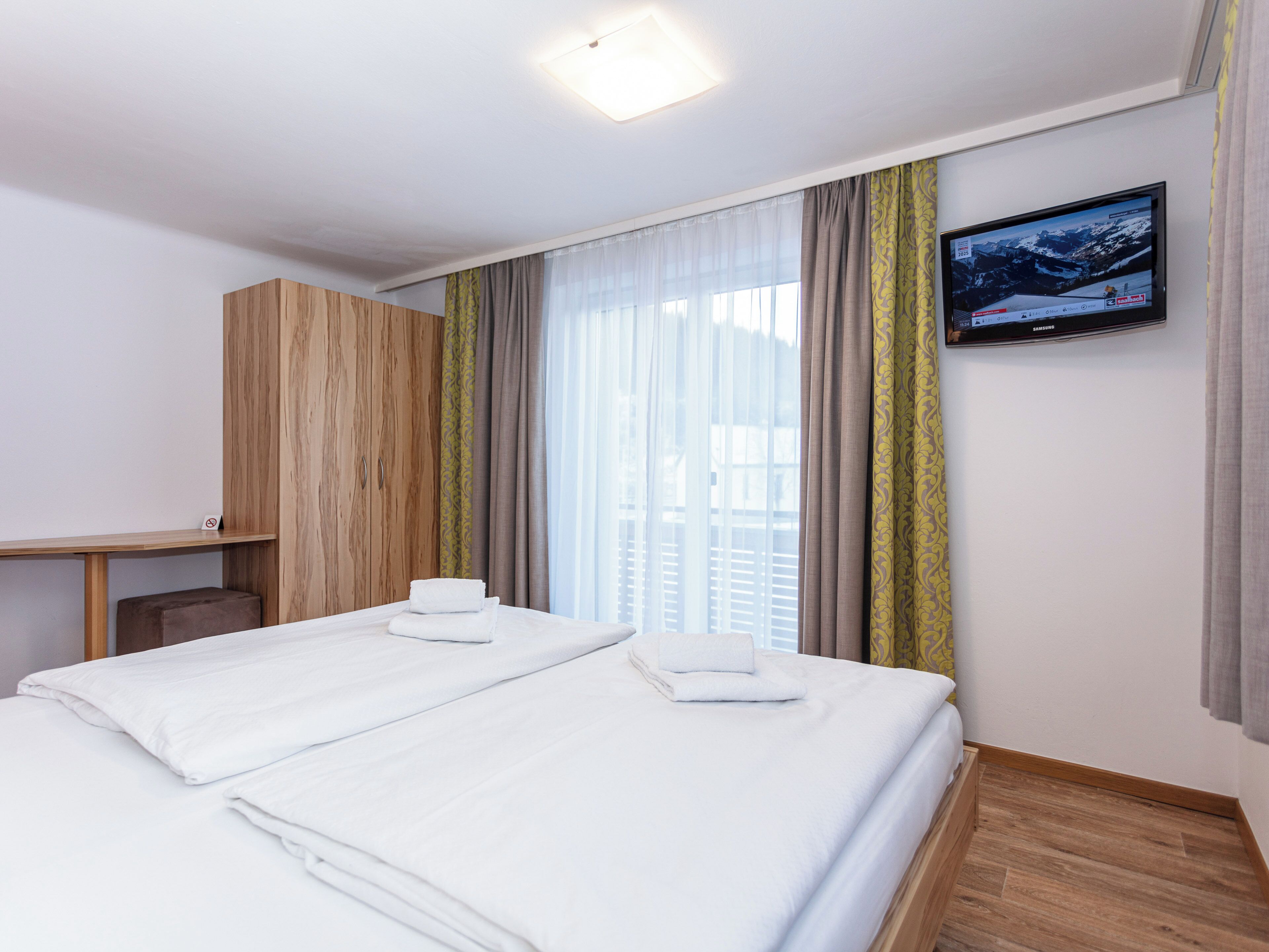 Habitación de hotel con dos camas, un armario de madera, un escritorio, un televisor en la pared y una ventana con cortinas.