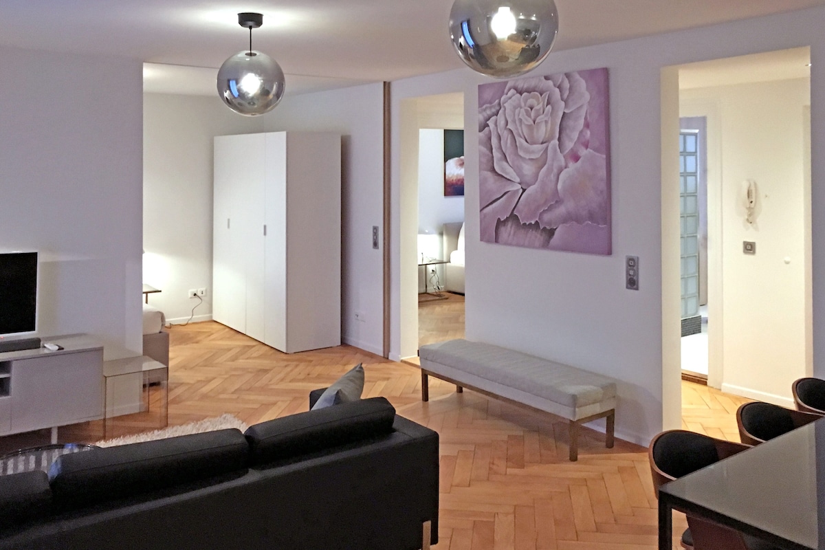 A modern living room with a sofa, a dining table, and chairs. There is a large artwork on the wall and a bench in the corner.