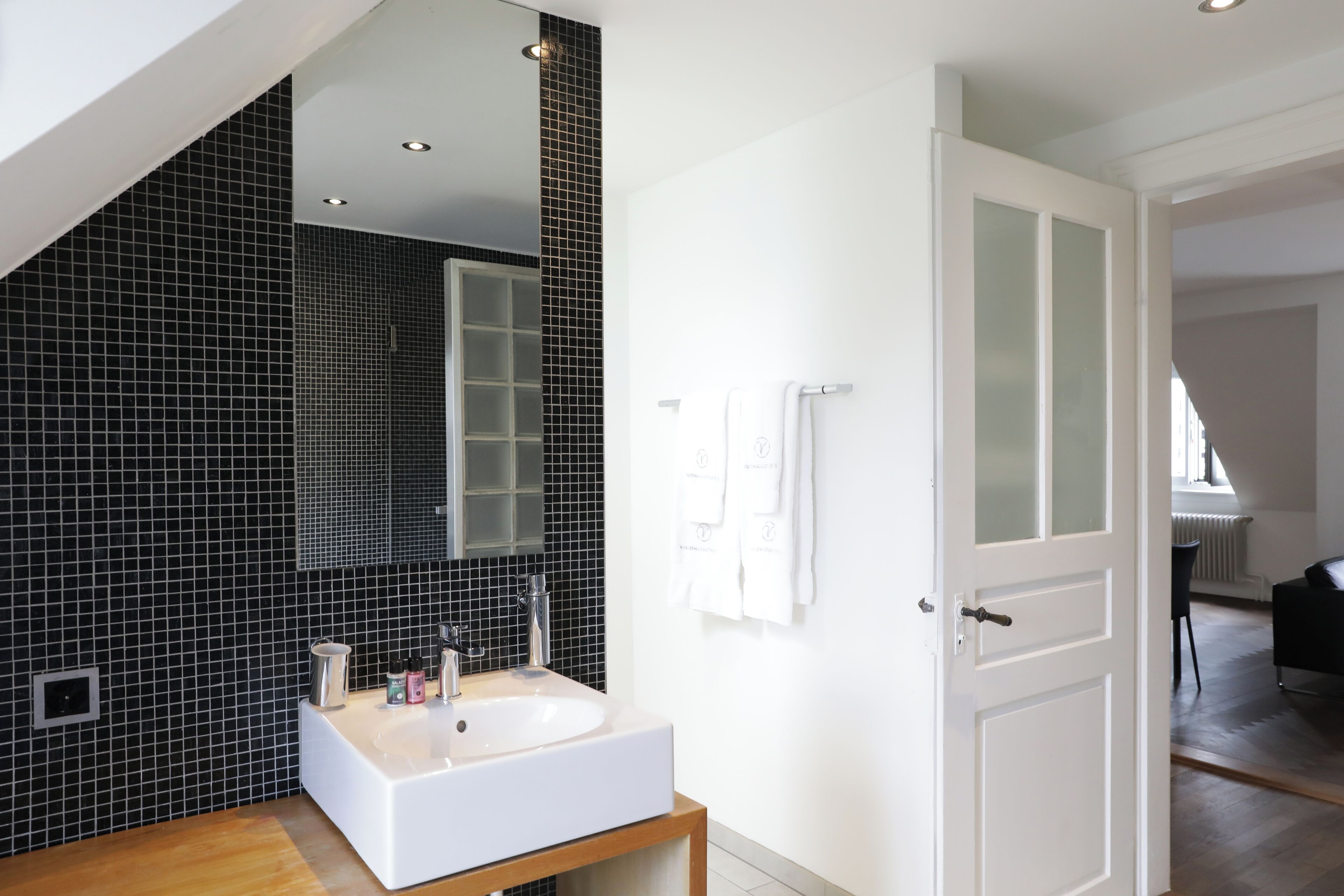 Un baño moderno con un lavamanos blanco, paredes revestidas de baldosas negras y una puerta que da a otra habitación.