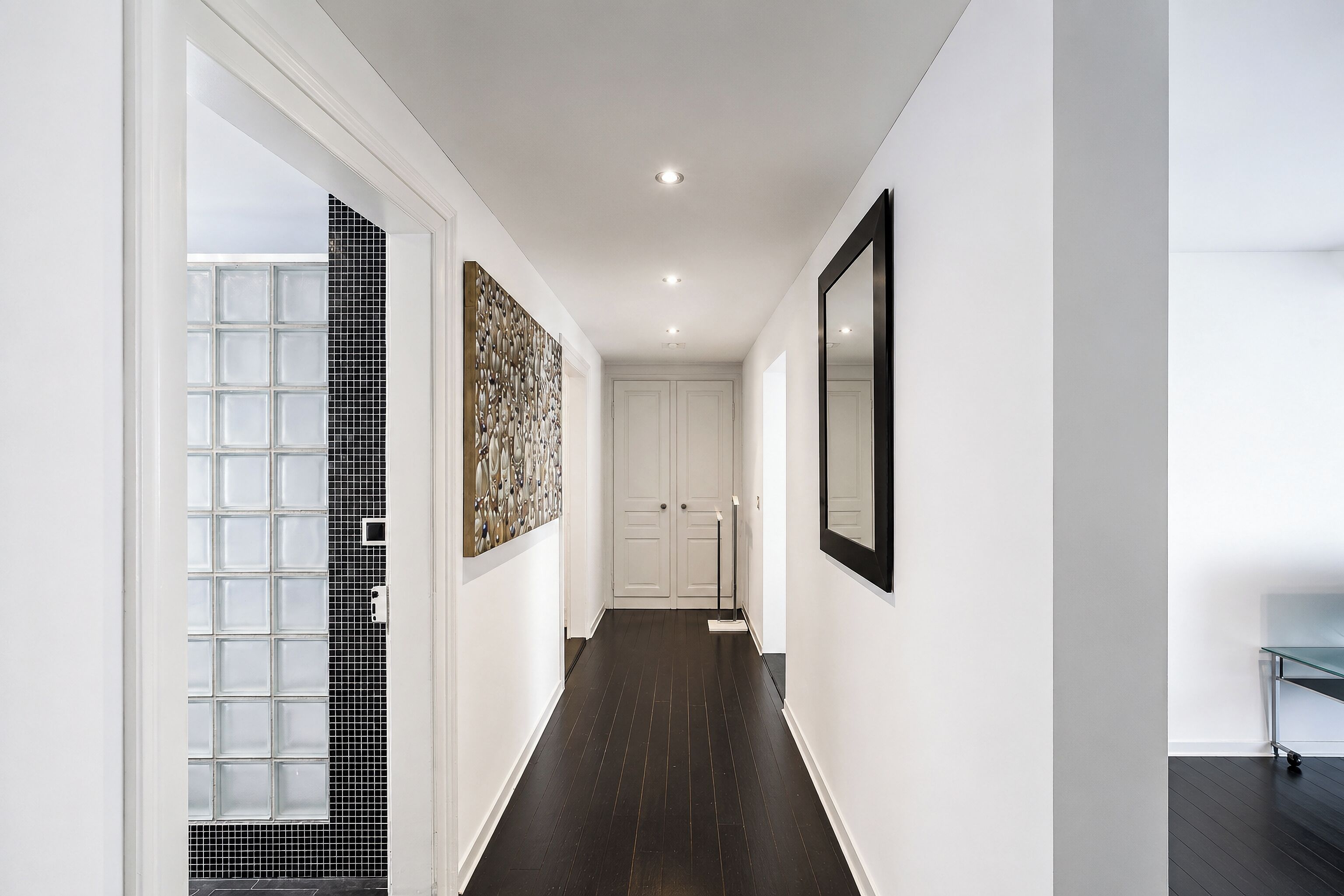 A hallway with a dark wooden floor, white walls, a large mirror, and a door at the end.