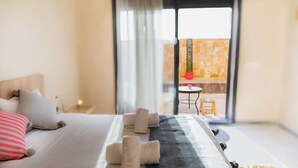 A hotel room with a bed, pillows, a blanket, and a set of towels. There is a view of an outdoor area with a table and chairs. - The Ocean Villa  (Dakhla)
