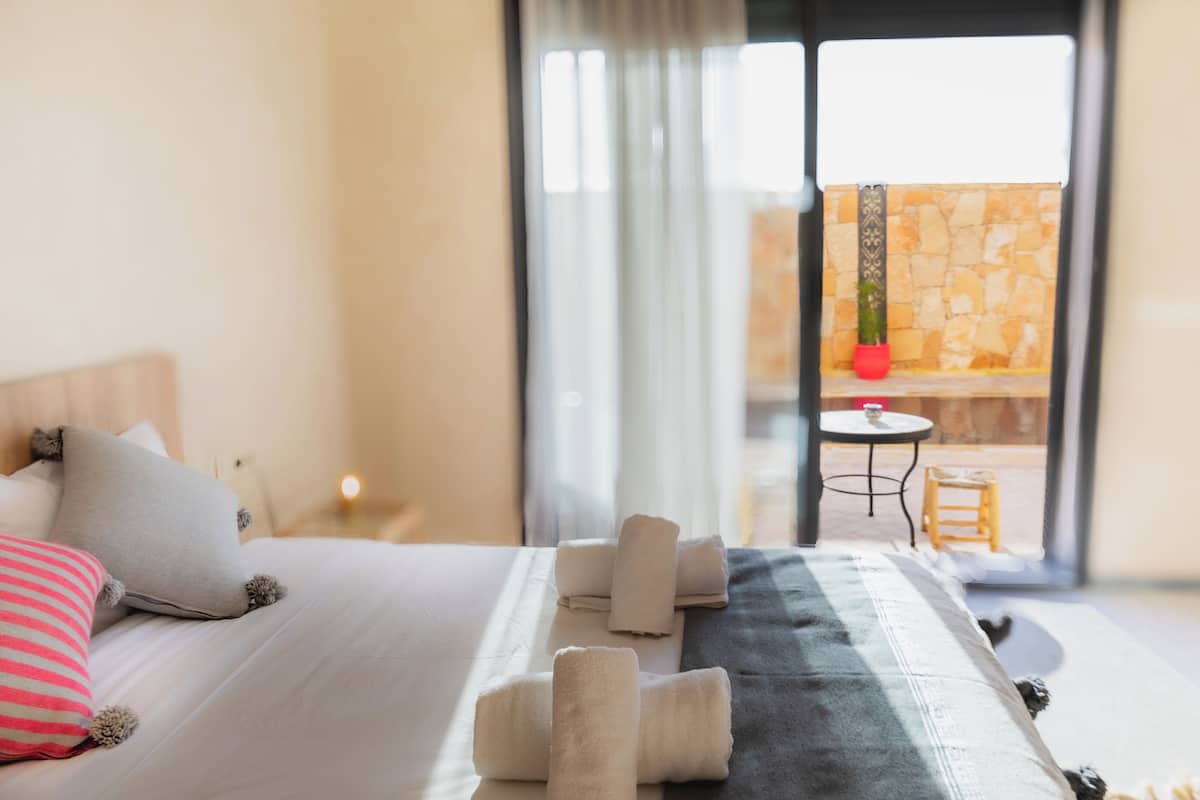 A hotel room with a bed, pillows, a blanket, and a set of towels. There is a view of an outdoor area with a table and chairs.
