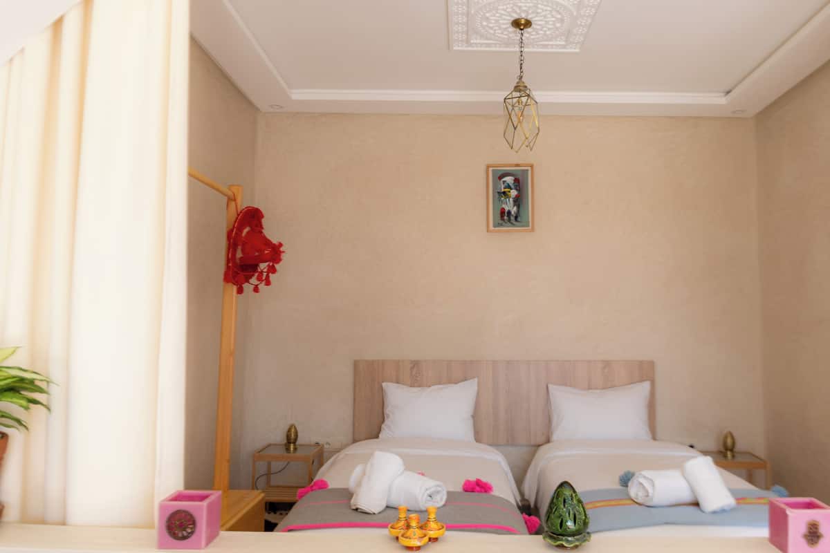 A hotel room with two beds, a wooden headboard, a ceiling light fixture, a framed picture on the wall, and a potted plant.