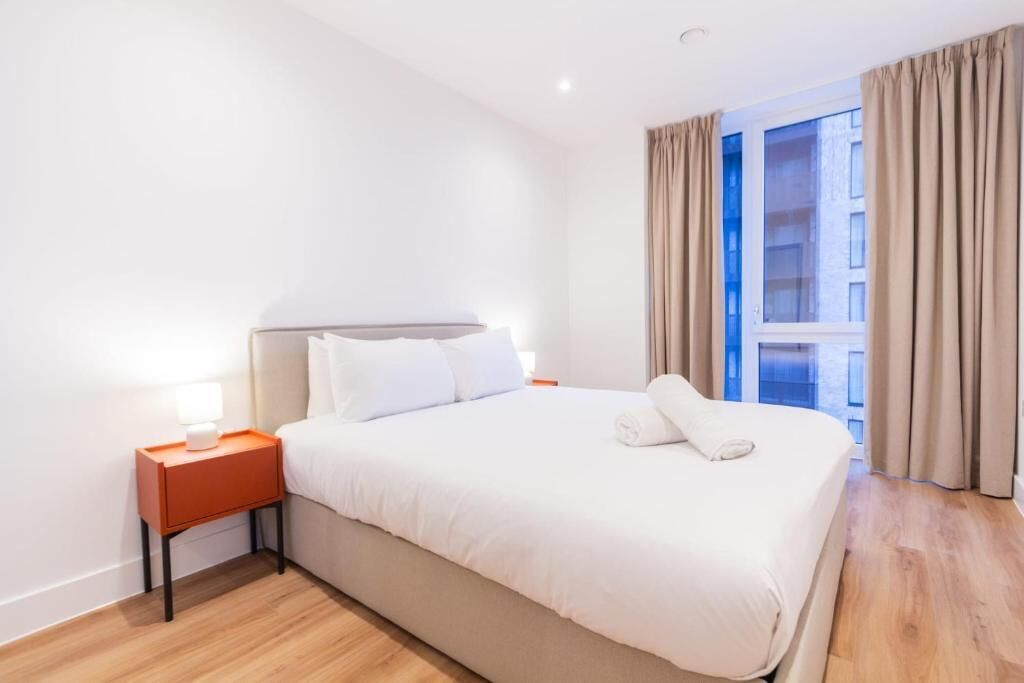 Ein Hotelzimmer mit einem großen Bett, einem Nachttisch mit Lampe und Blick auf die Stadt durch das Fenster.