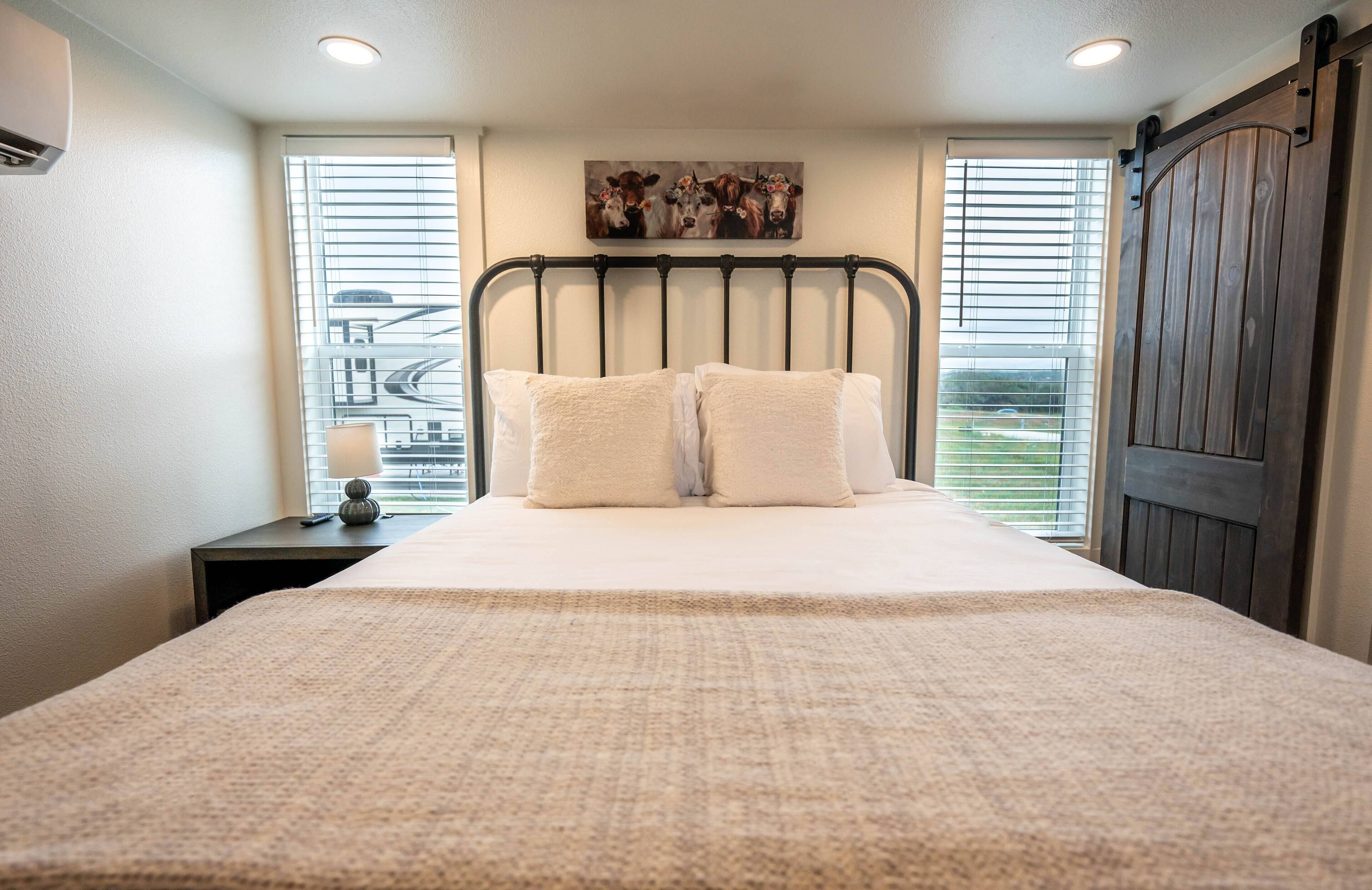 A bedroom with a large bed, two bedside tables, a window with blinds, a painting on the wall, and a wooden door.