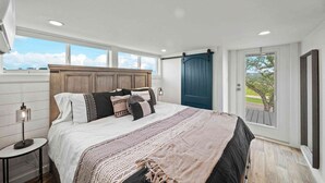 A bedroom with a large bed, a nightstand, a lamp, a window, and a door leading outside. - The Charming Chayil Cottage - 046 (Fredericksburg)