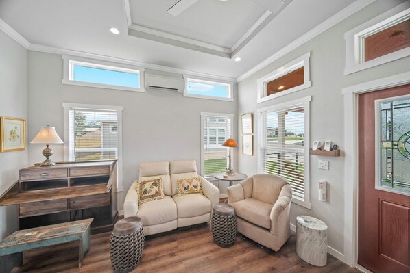 A living room with a sofa, a coffee table, a lamp, a bench, and a small side table. There are two windows with blinds, a door, and a ceiling with recessed lighting.