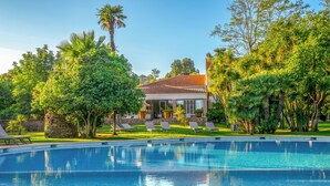 A swimming pool with lounge chairs, surrounded by lush greenery and a building with a tiled roof. - Château de Valmer (La Croix-Valmer)
