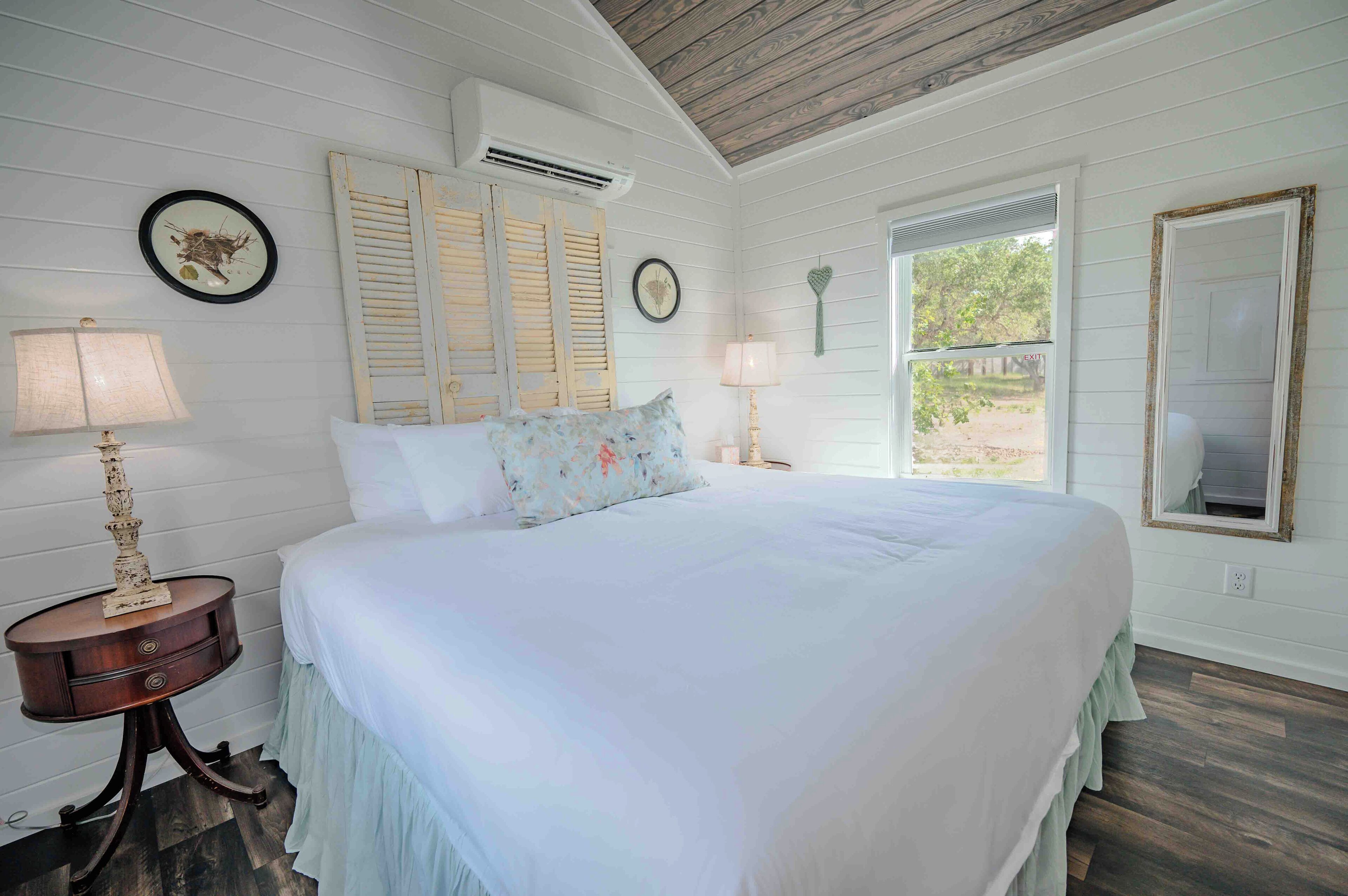 A bedroom with a large bed, two bedside tables with lamps, a window with shutters, and a mirror on the wall.