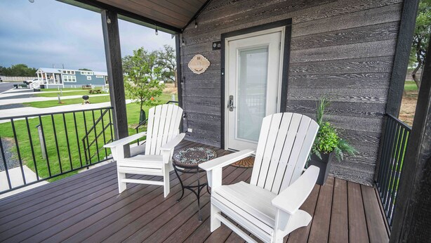 A covered porch with two white Adirondack chairs and a small table, overlooking a grassy area and a building in the background.