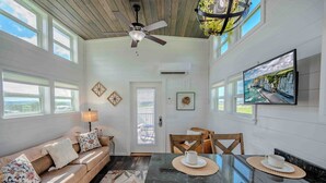 A spacious living area with a sofa, dining table, and chairs. A ceiling fan and TV are mounted on the wall.