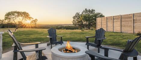 A circular fire pit with four Adirondack chairs around it, set on a concrete patio with a view of a grassy field and a wooden fence in the background.