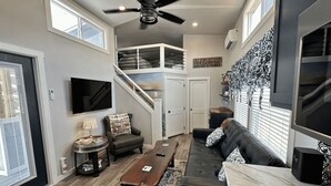 A spacious living room with a high ceiling fan, a staircase leading to an upper level, a black leather sofa, a wooden coffee table, and a TV mounted on the wall.
