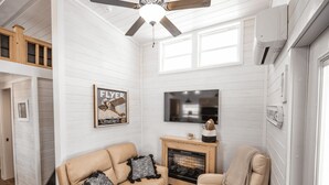 Un salon avec un canapé beige, une cheminée, un téléviseur et un ventilateur de plafond.