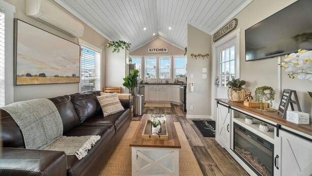 A modern living room with a brown leather sofa, a wooden coffee table, and a kitchen in the background. - The Haven - 95 (Fredericksburg)