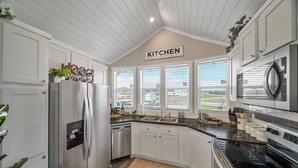 A modern kitchen with white cabinets, stainless steel appliances, a built-in oven, and a large window with blinds. - The Haven - 95 (Fredericksburg)