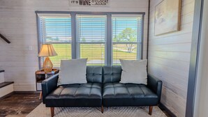 A dark leather sofa with two white cushions, a lamp, and a sign above the windows that reads "It's WINE O'CLOCK...AGAIN".