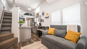 A modern living space with a staircase, a grey sofa with yellow cushions, a kitchen area, and a dining table. - Cottage on the Hill - 114 (Fredericksburg)