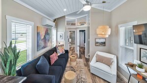 A modern living room with a dark blue sofa, a white armchair, and a large TV. There is a ceiling fan, a woven rug, and a staircase leading to an upper level. - Comfortably Numb - 119 (Fredericksburg)