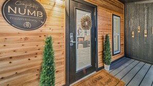 A wooden exterior with a black door, a welcome mat, and a sign reading "Comfortably NUMB Fredericksburg, TX". - Comfortably Numb - 119 (Fredericksburg)