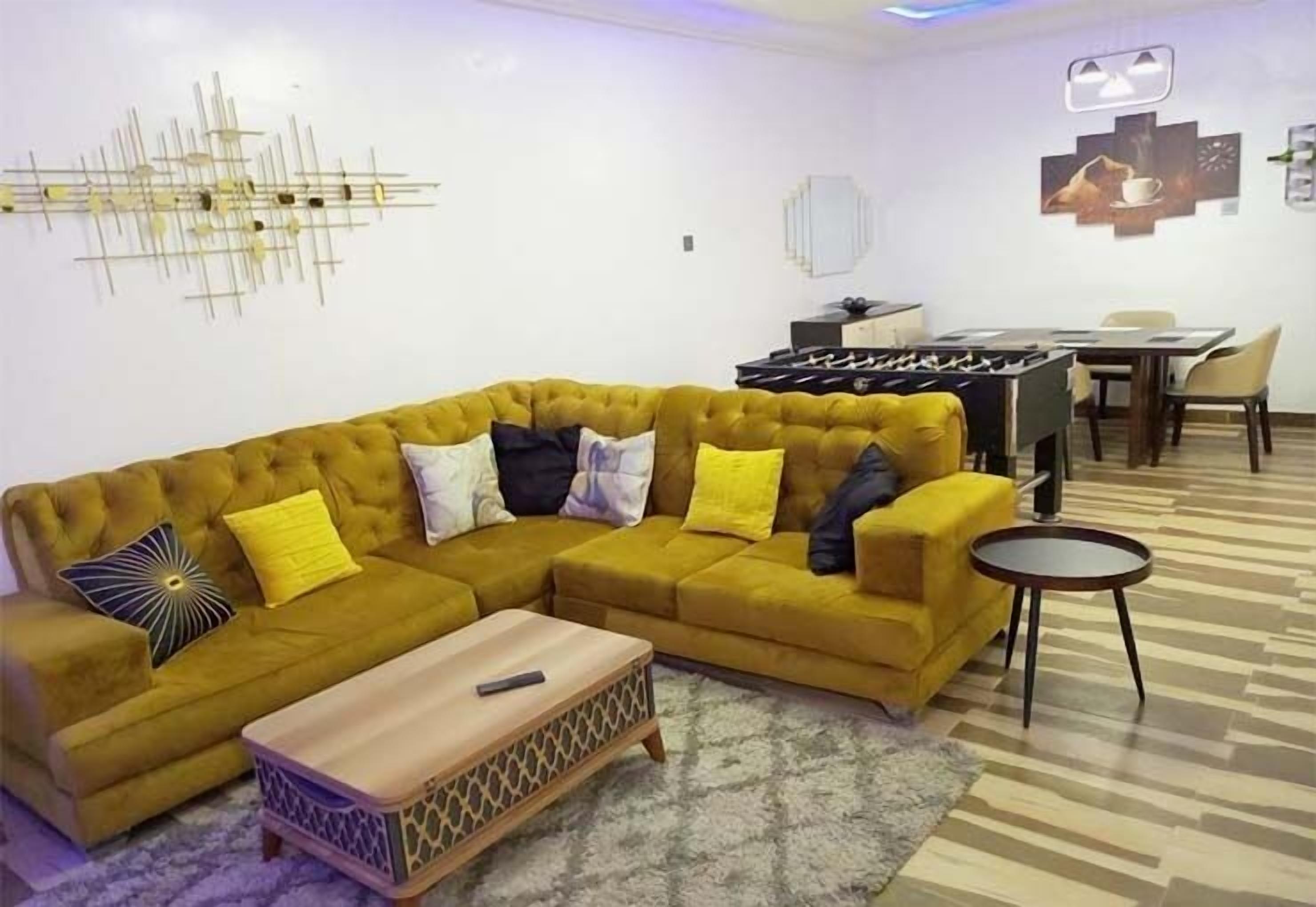 A living room with a mustard-colored sofa, a wooden coffee table, a patterned rug, a foosball table, and a dining area with chairs and a table.