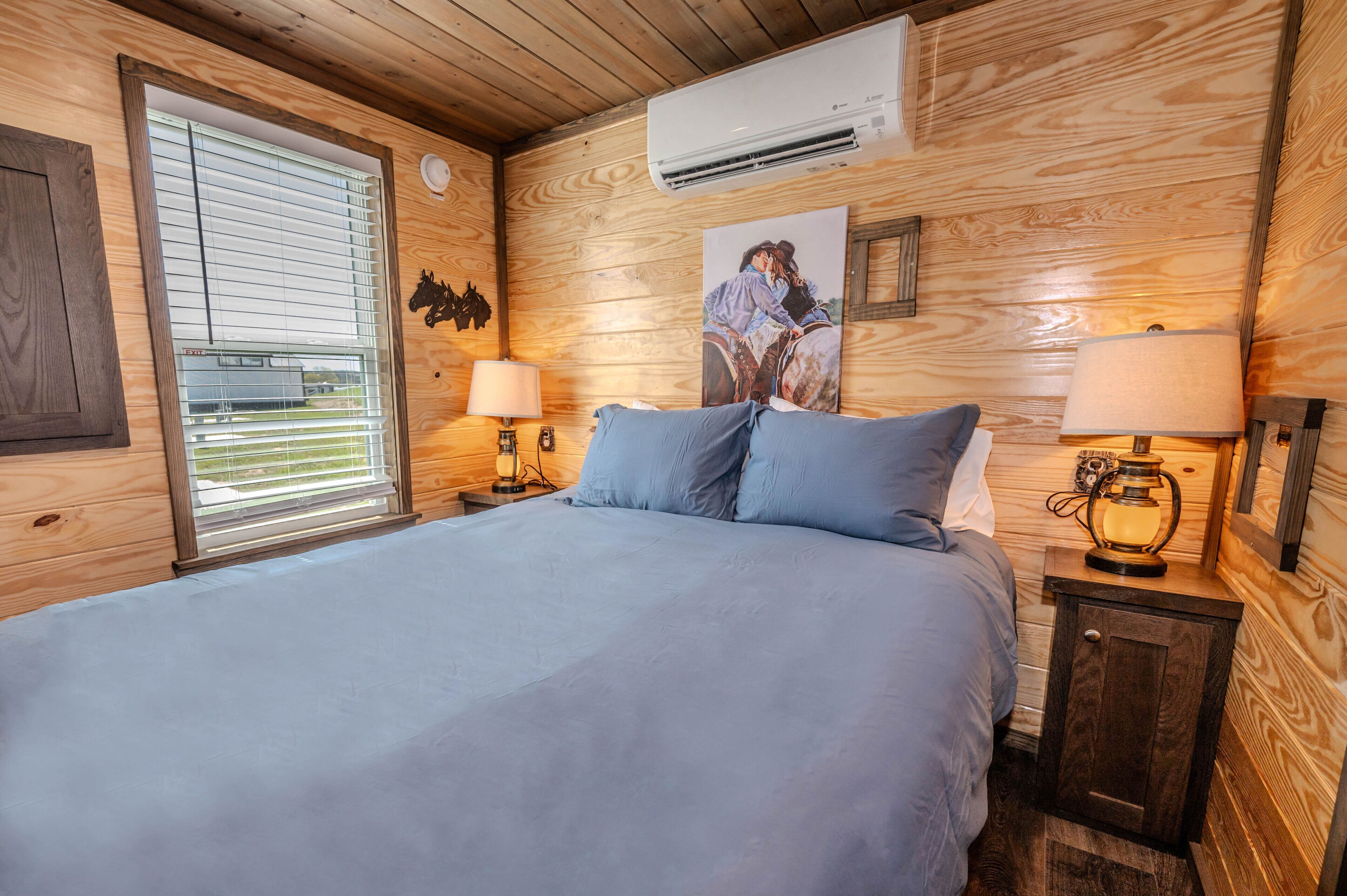 Ein Schlafzimmer einer Holzhütte mit einem Bett, zwei Nachttischlampen, einer Klimaanlage und einem Fenster mit Jalousien.