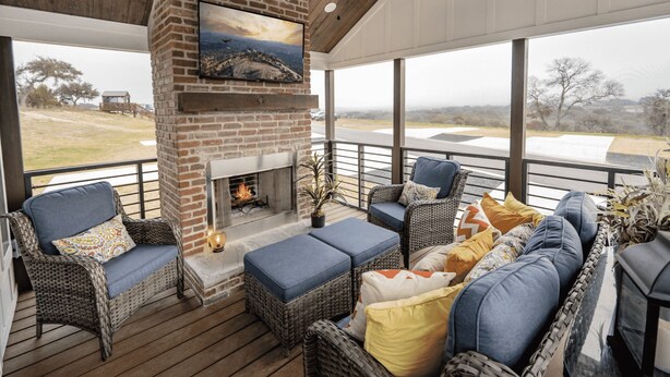 A covered outdoor seating area with wicker furniture, a fireplace, and a television mounted on a brick wall. - The Little Haus - 153 (Fredericksburg)