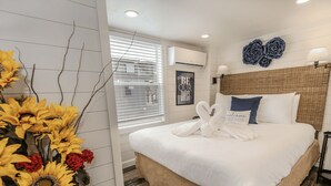 A hotel room with a large bed, a wall-mounted air conditioner, a framed 'BE OUR GUEST' sign, and a decorative wall piece. - The Little Haus - 153 (Fredericksburg)