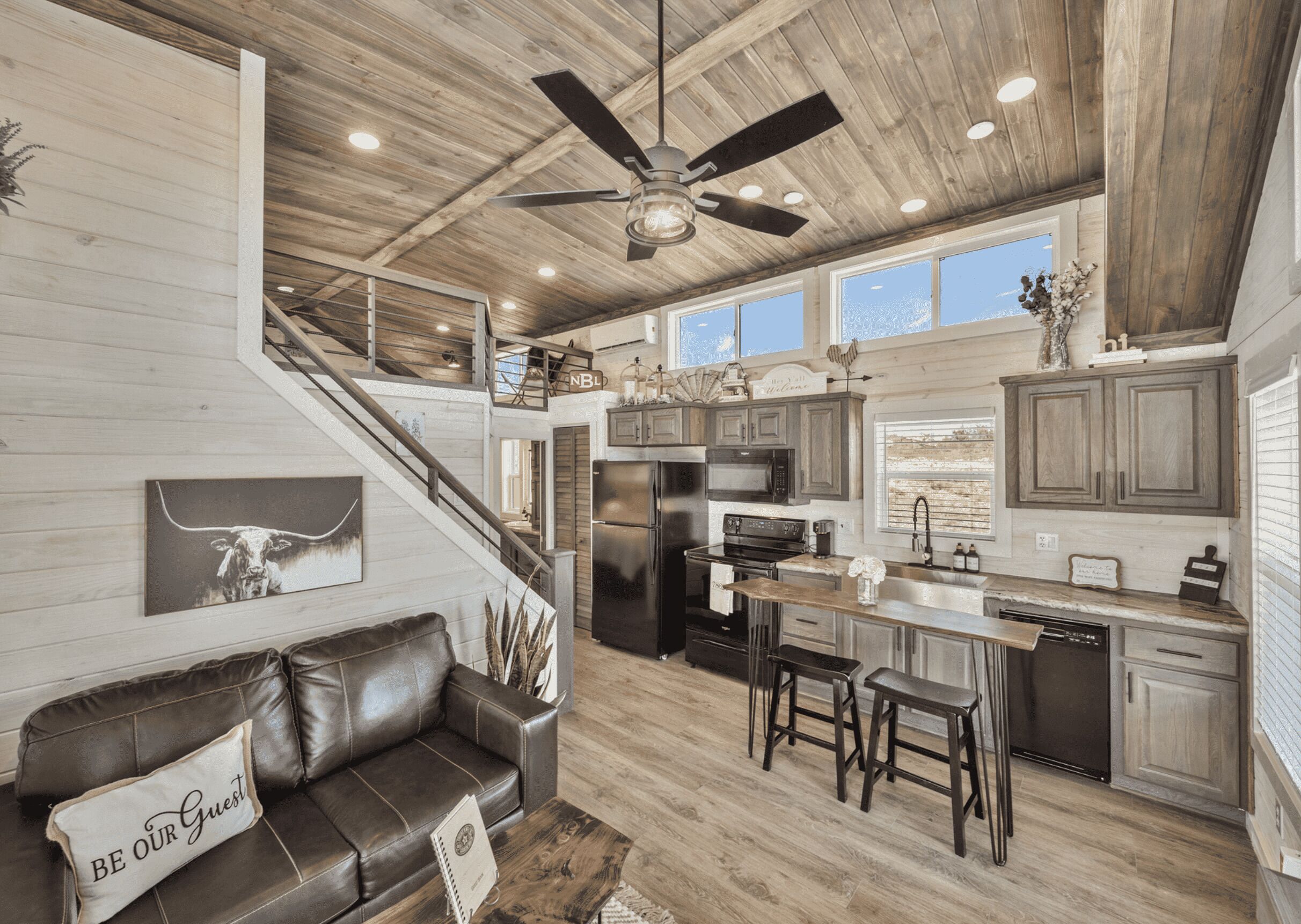 A spacious kitchen with a central island, a dining area with bar stools, a leather sofa with a 'BE OUR GUEST' pillow, and a staircase leading to a loft area.