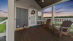 Un porche cubierto con suelo de madera, dos sillas Adirondack, una puerta decorada con un adorno y vistas del paisaje.