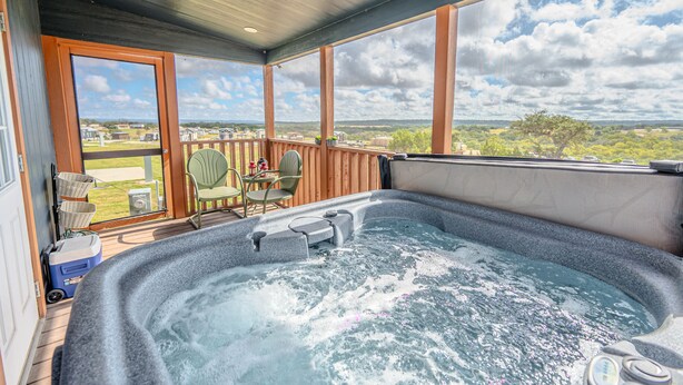 A hot tub with a view of a grassy area and trees, a wooden deck, and a covered patio area with chairs and a table.